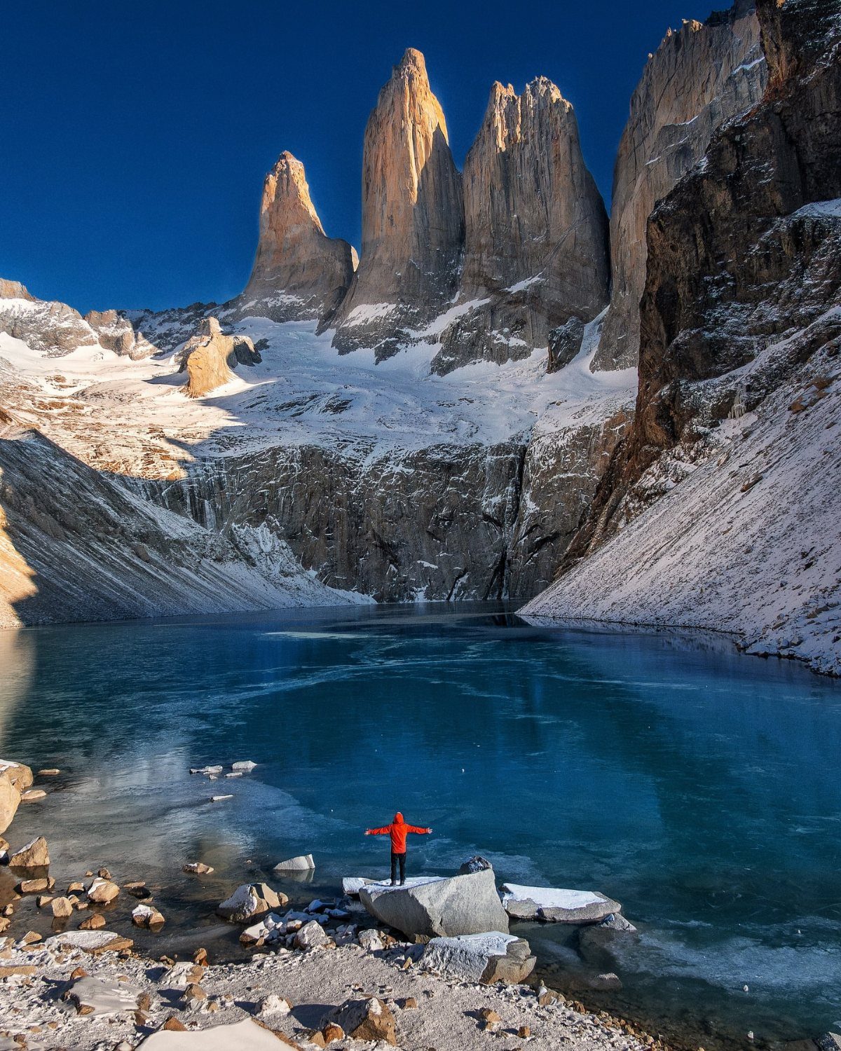 Person in red jacket facing towering granite peaks and a turquoise glacial lake.