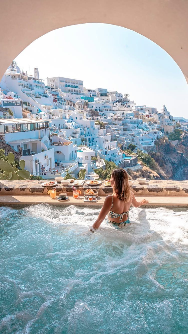 Woman in a pool overlooking the whitewashed buildings of Santorini through an archway