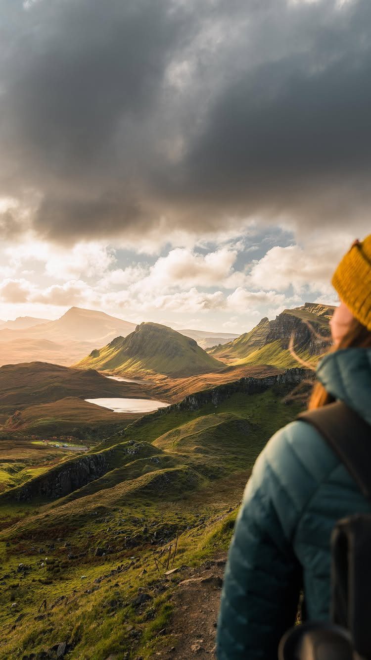 16 Adventurous Places to Travel, From Utah to Mongolia 13 Hiker in yellow beanie overlooking green Scottish highlands and distant lakes under clouds.