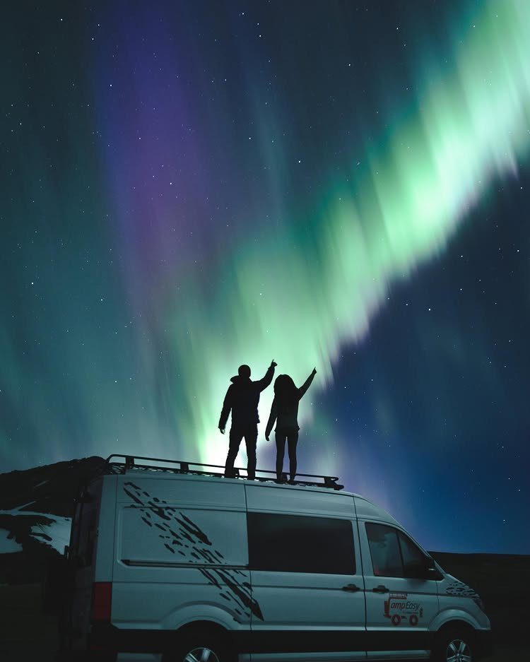 Couple standing on a camper van roof under a green aurora borealis sky.