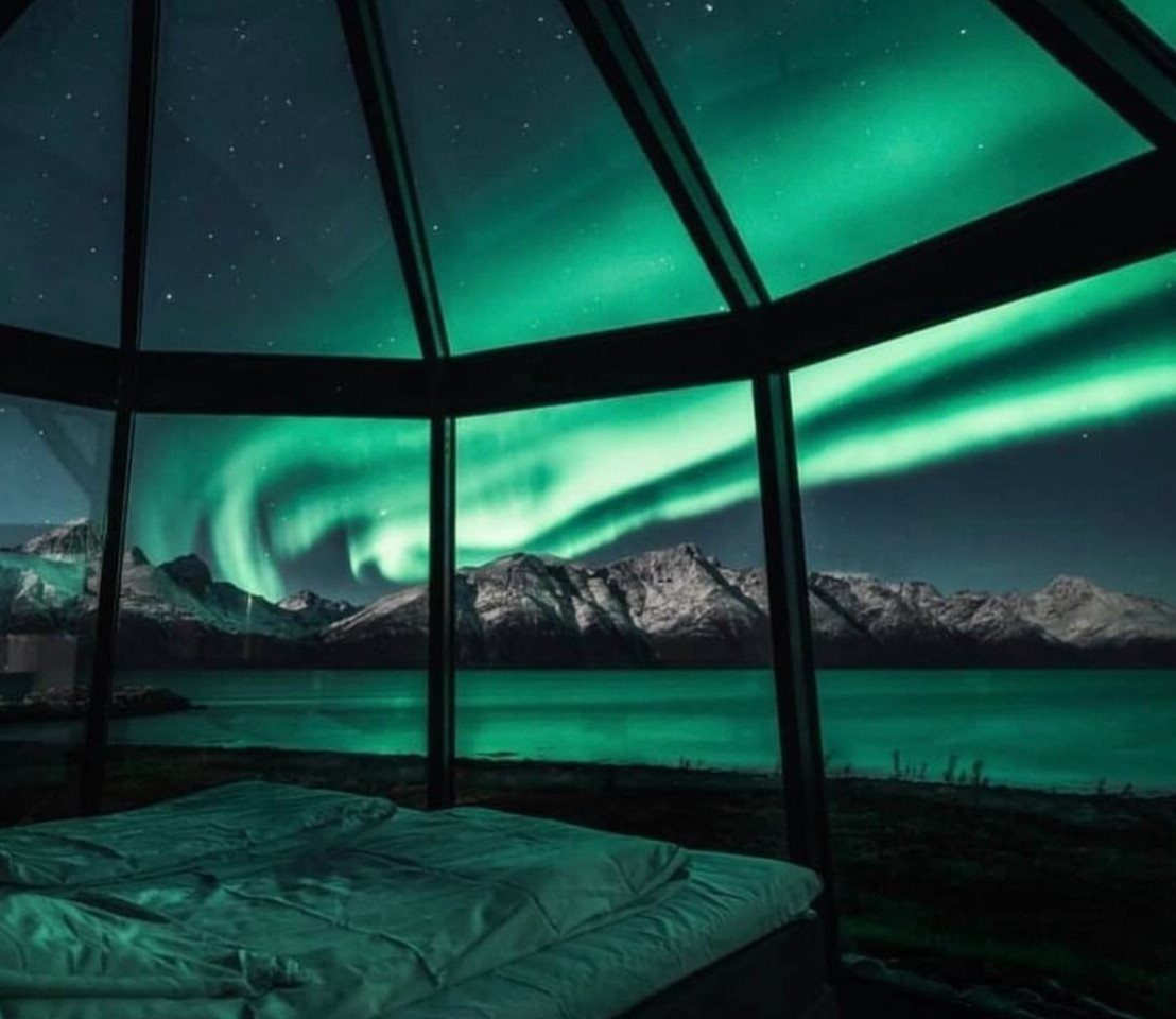 Glass igloo interior with a bed overlooking green northern lights and snowy mountains.