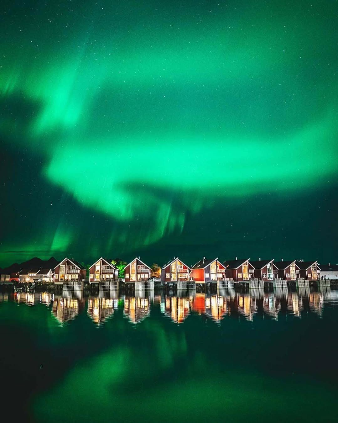 Green aurora borealis shimmering over a row of waterfront houses at night.