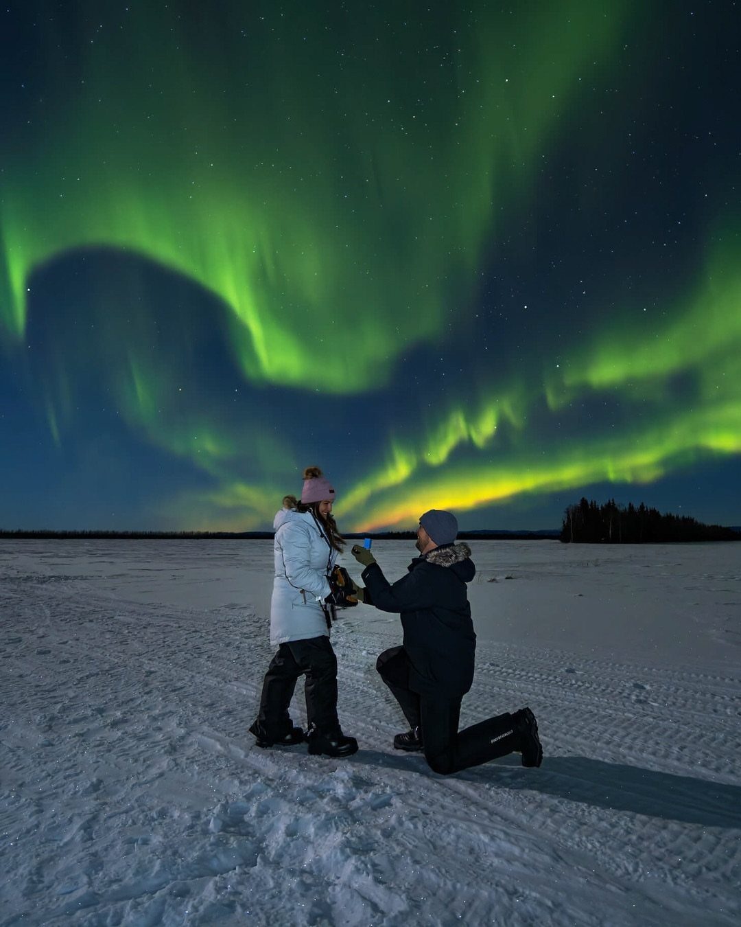 Couple proposing on a snowy field under a vibrant green aurora borealis sky