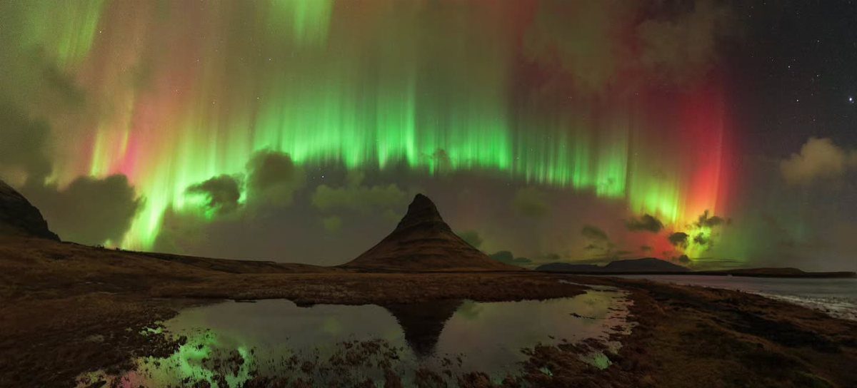 Green and red northern lights over a pointed mountain reflected in water.