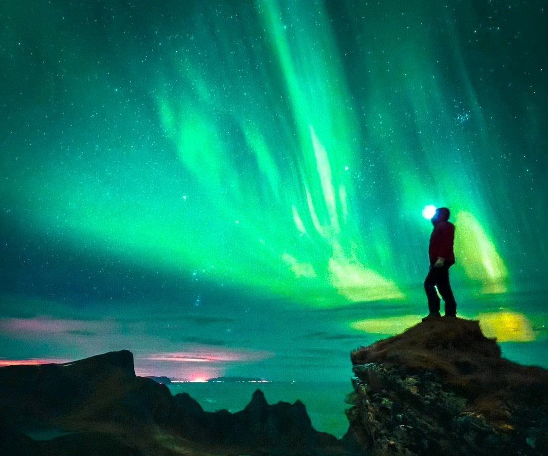 Person standing on a rocky cliff under a bright green aurora borealis sky