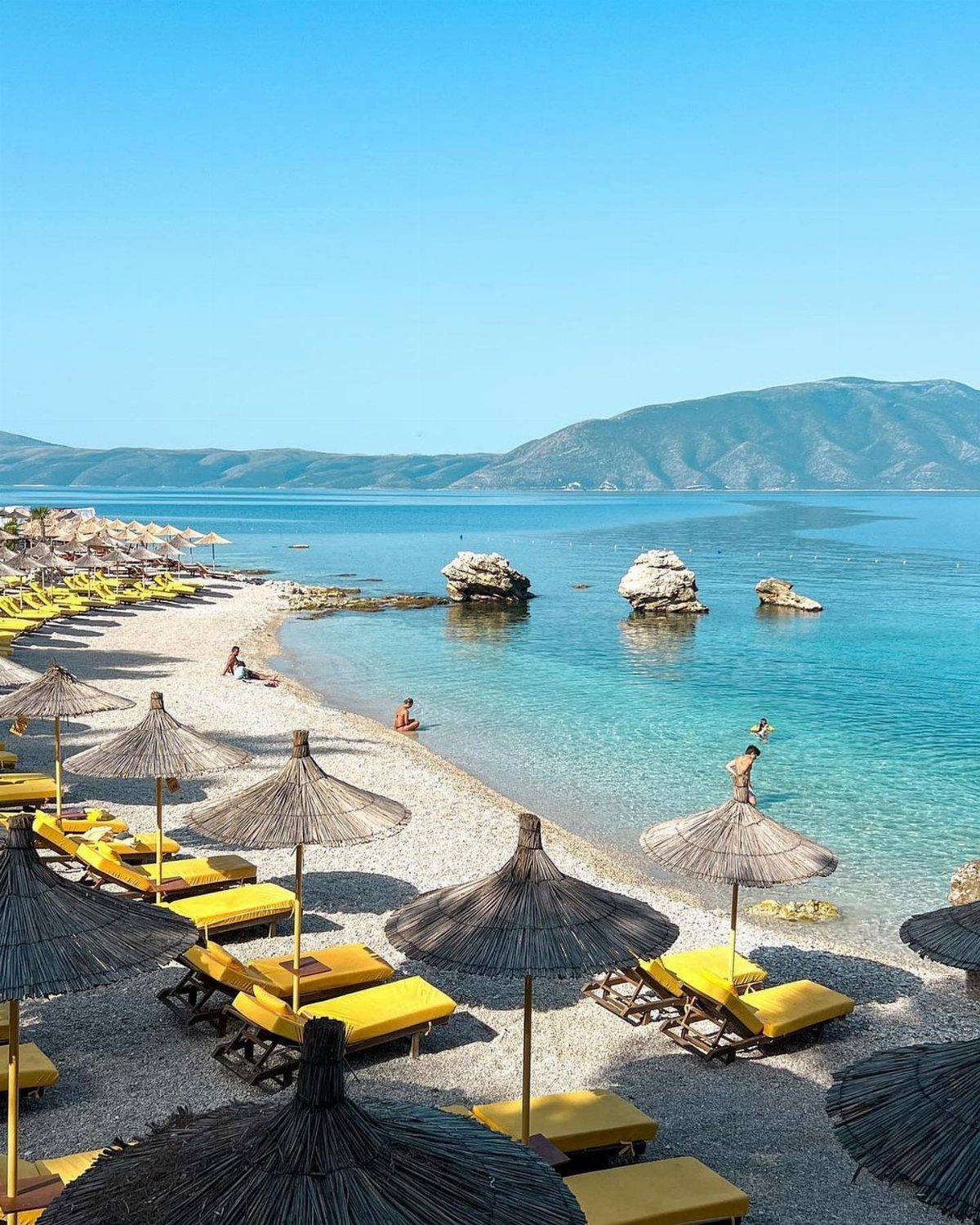 Beach Places to Travel: 13 Ideas for Your Next Trip 13 Turquoise beach with yellow lounge chairs, thatched umbrellas, and distant blue mountains.