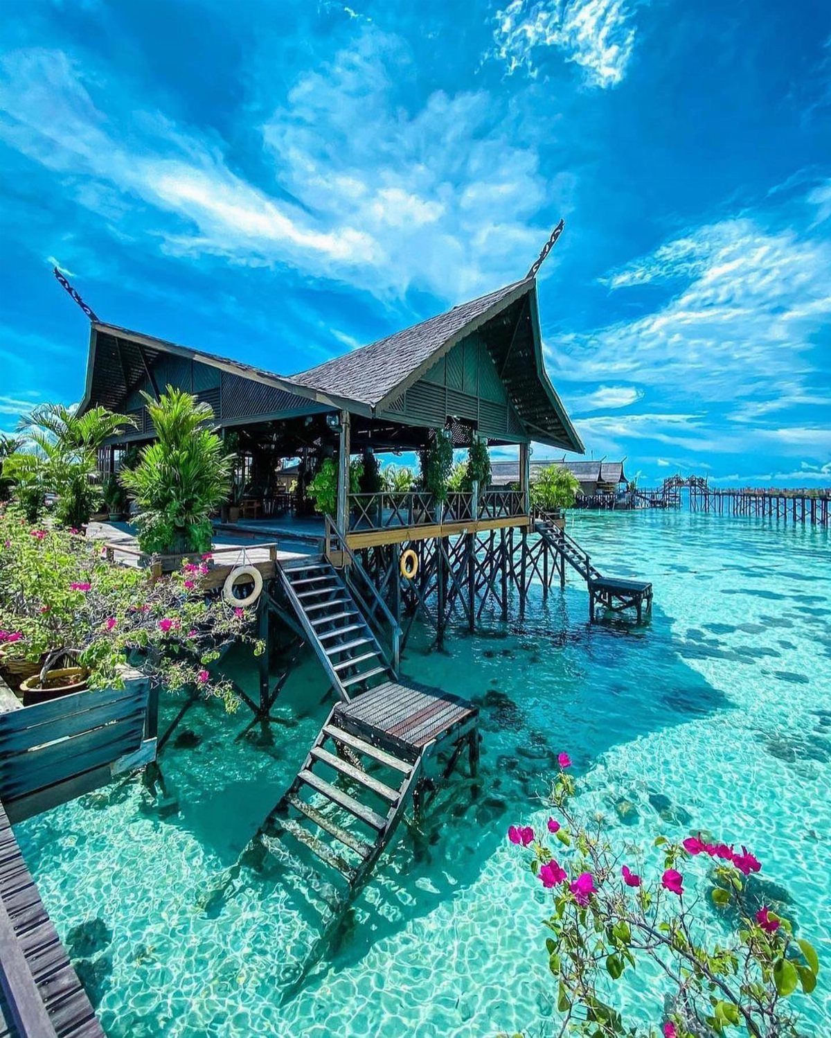 Beach Places to Travel: 13 Ideas for Your Next Trip 12 Wooden stilt house over clear turquoise water with tropical plants and blue sky.