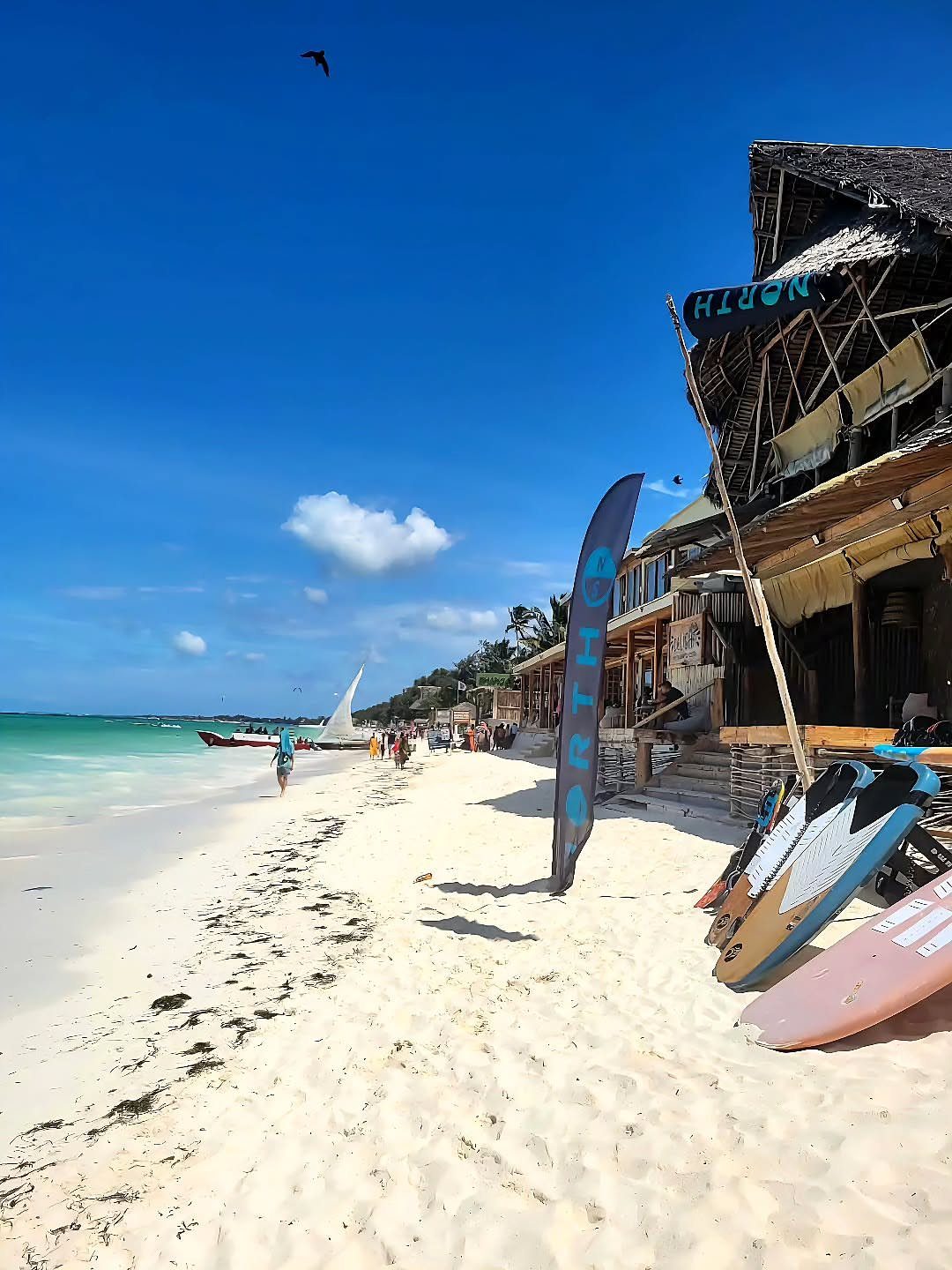Beach Places to Travel: 13 Ideas for Your Next Trip 7 White sandy beach with turquoise water, surfboards, and a thatched roof building.