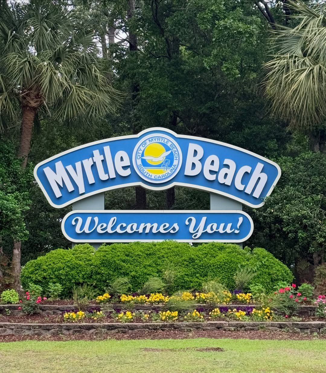 6 Beach Places to Visit, From Cape May to the Kona Coast 2 Myrtle Beach welcome sign surrounded by green shrubbery, flowers, and palm trees.