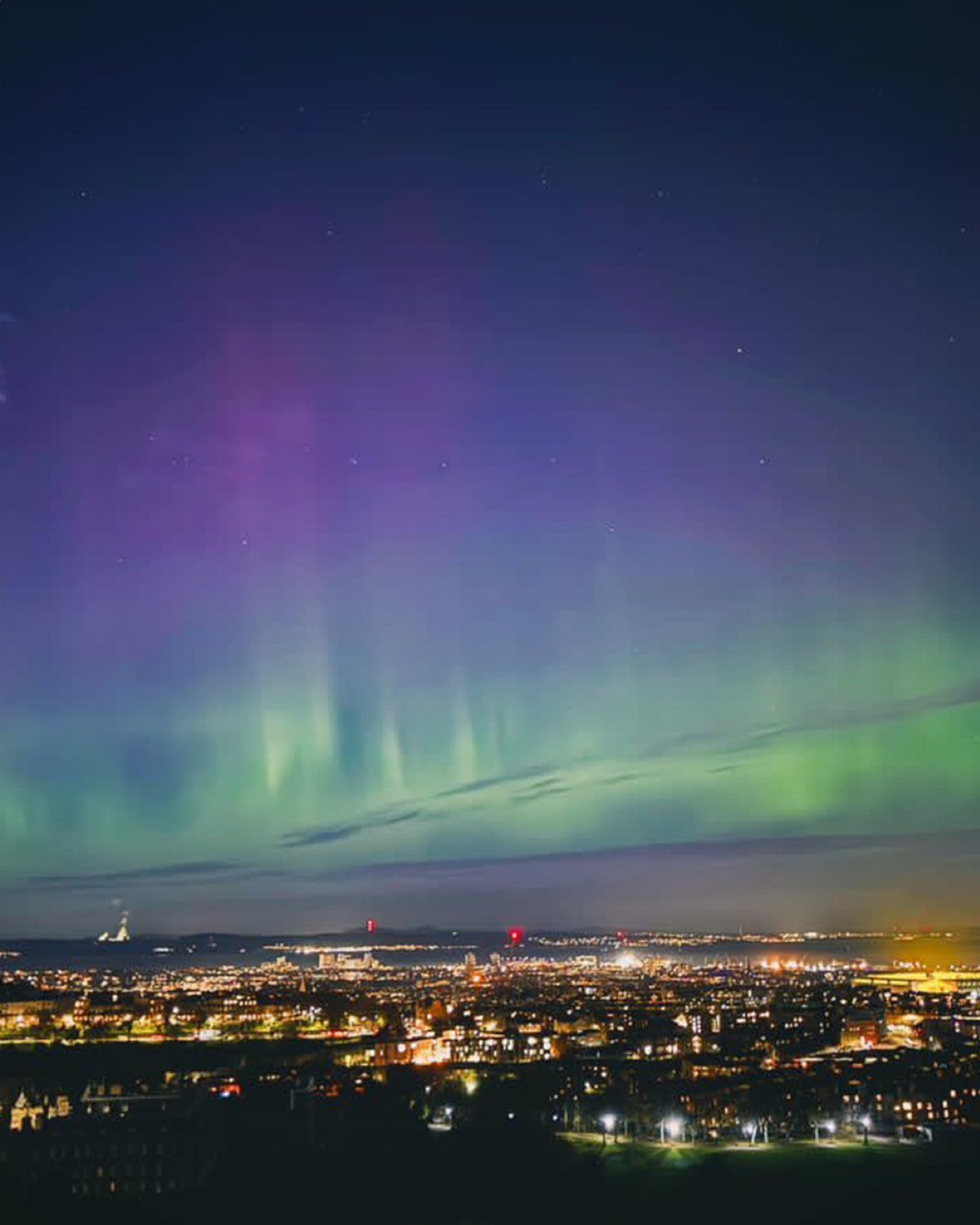 Aurora borealis with green and purple lights over a glowing night city skyline.