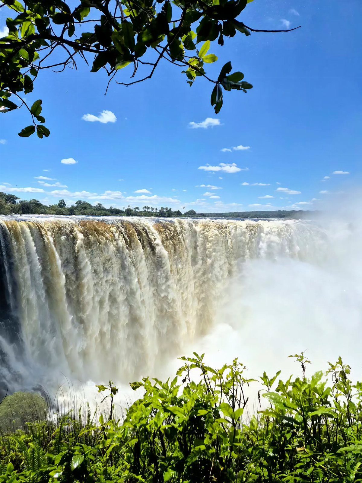 Wide waterfall cascading over a cliff into mist under a blue sky.