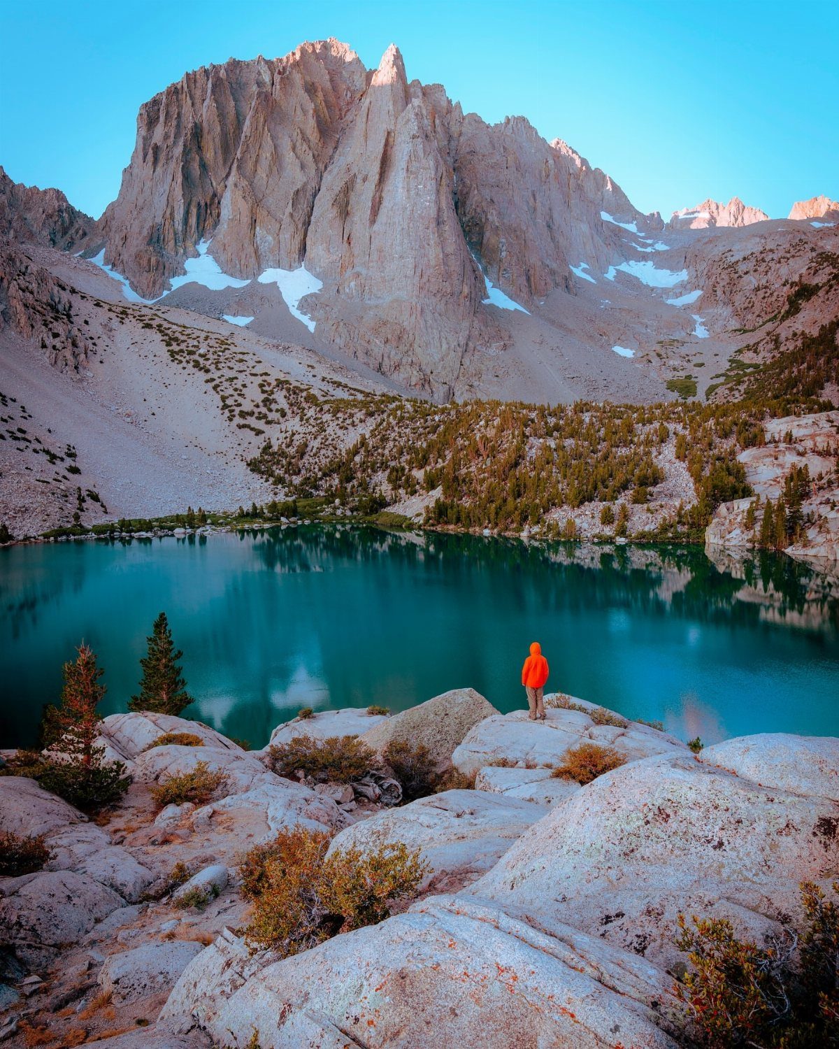 Person in orange jacket overlooking a turquoise lake and jagged granite mountain peaks.