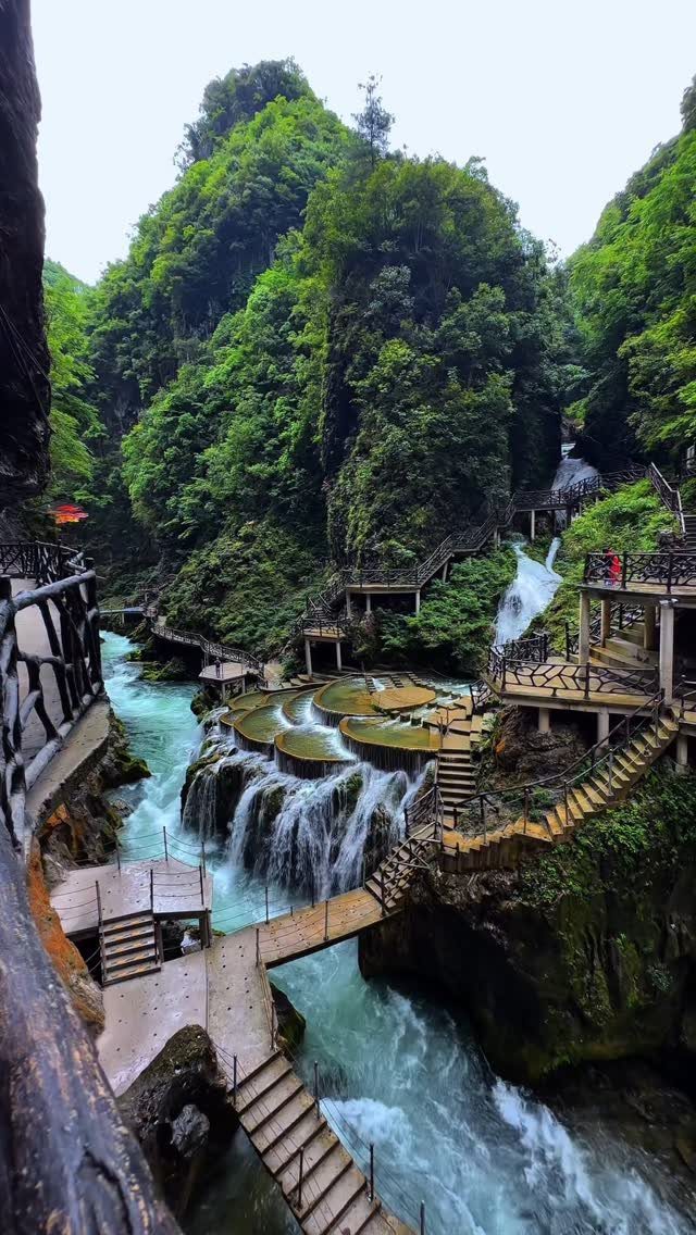 Lush green canyon with cascading waterfalls and wooden walkways over turquoise water.