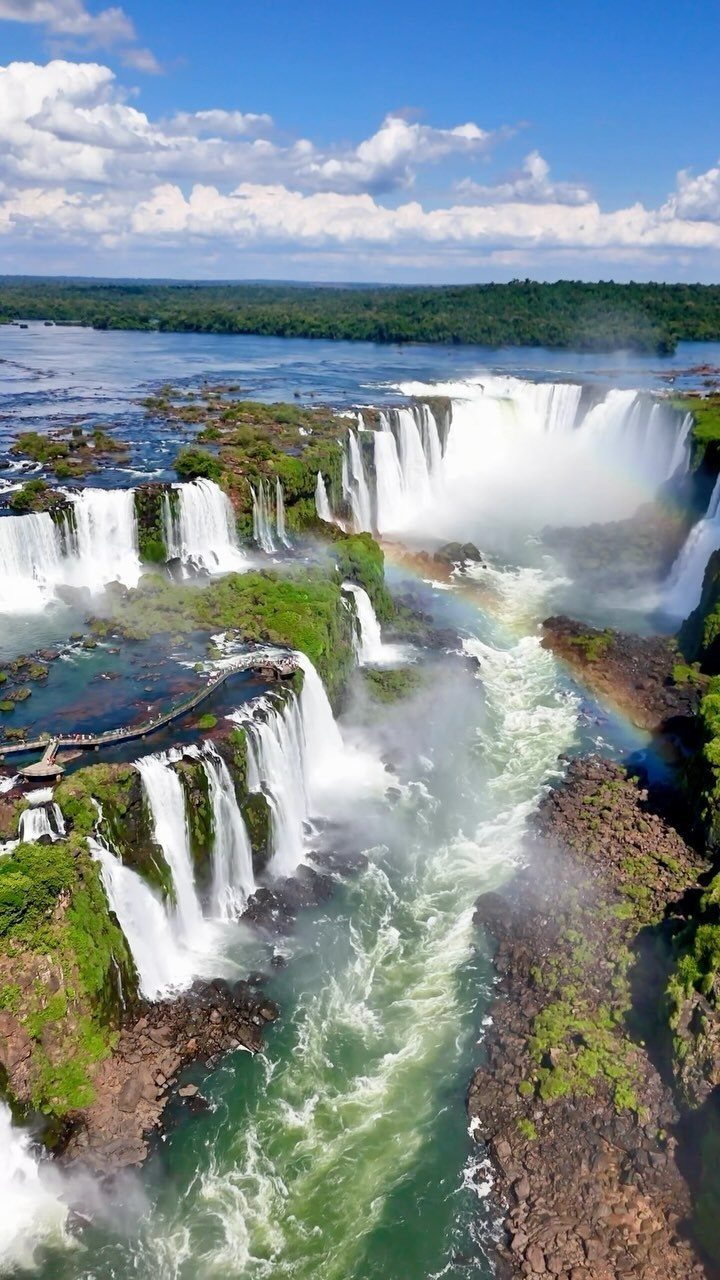Massive waterfalls with lush green cliffs and a rainbow under a blue sky.
