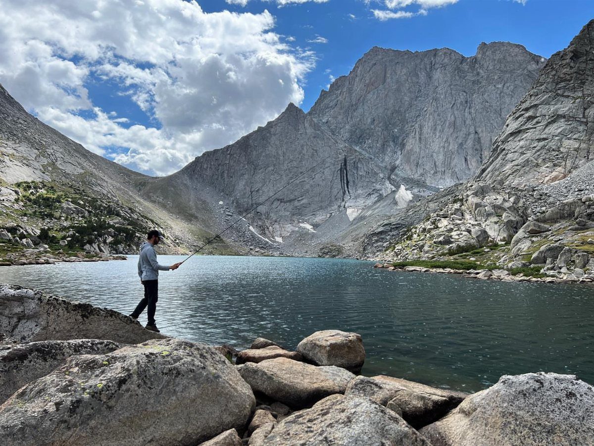 Person fishing at a blue alpine lake surrounded by rugged grey mountain peaks