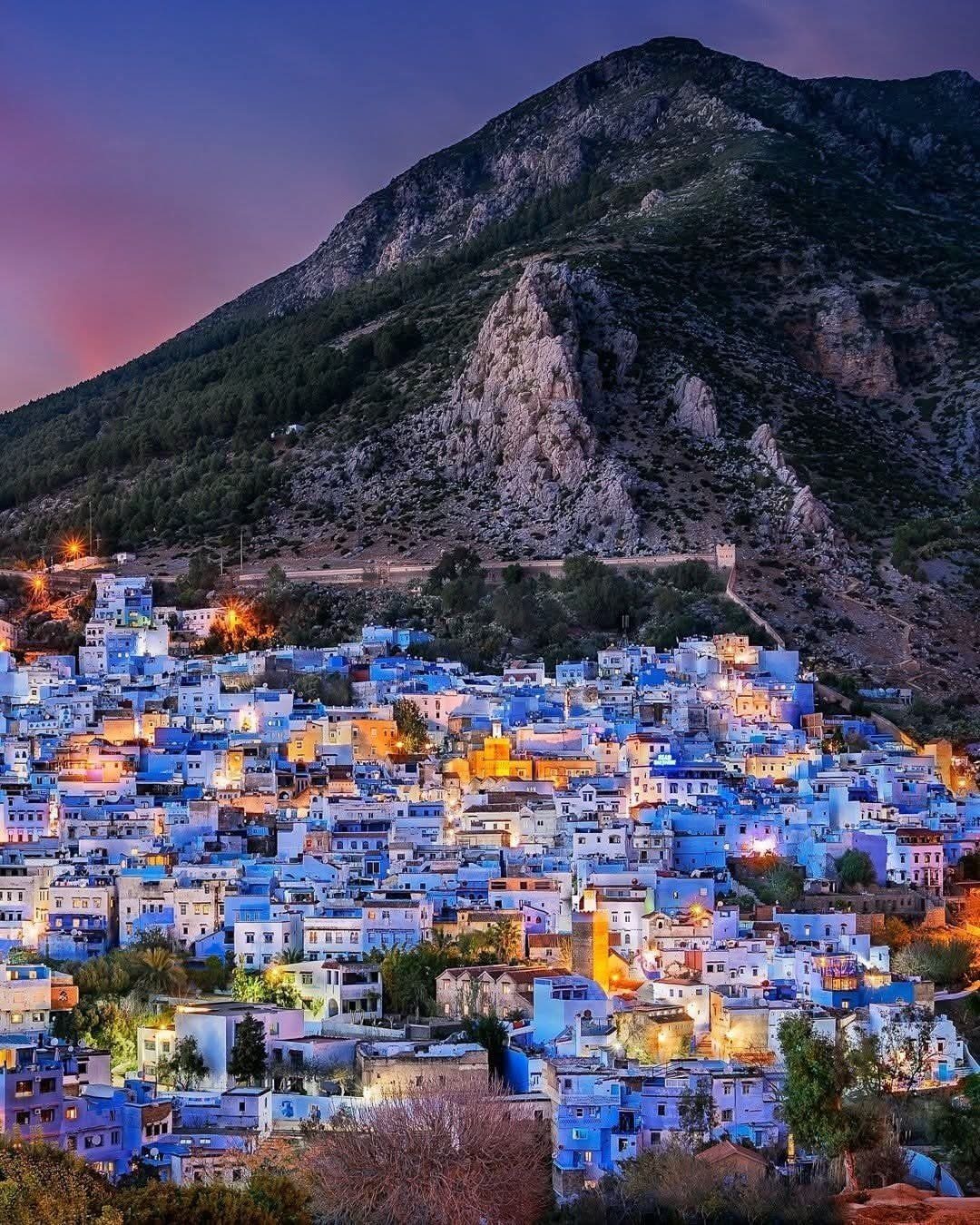 28 Beautiful Places to Travel Around the World 22 Blue buildings of a hillside town nestled beneath a large rocky mountain at twilight.