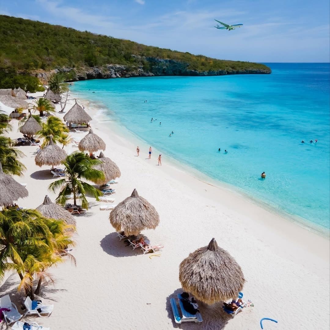 28 Beautiful Places to Travel Around the World 10 Tropical beach with white sand, turquoise water, thatched umbrellas, and a flying plane.