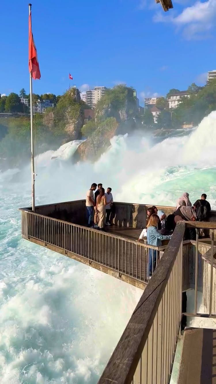 Bucket List Switzerland: 13 Scenic Places and Experiences 5 People on a viewing platform overlooking the powerful Rhine Falls in Switzerland.