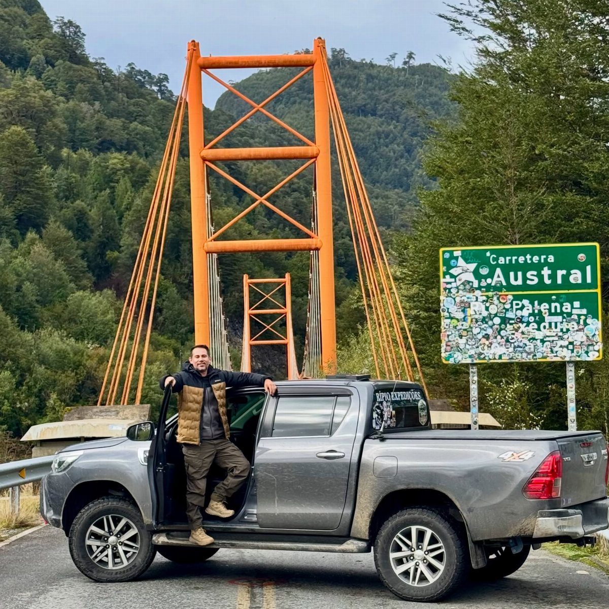 Man and grey pickup truck by a Carretera Austral sign and orange bridge.