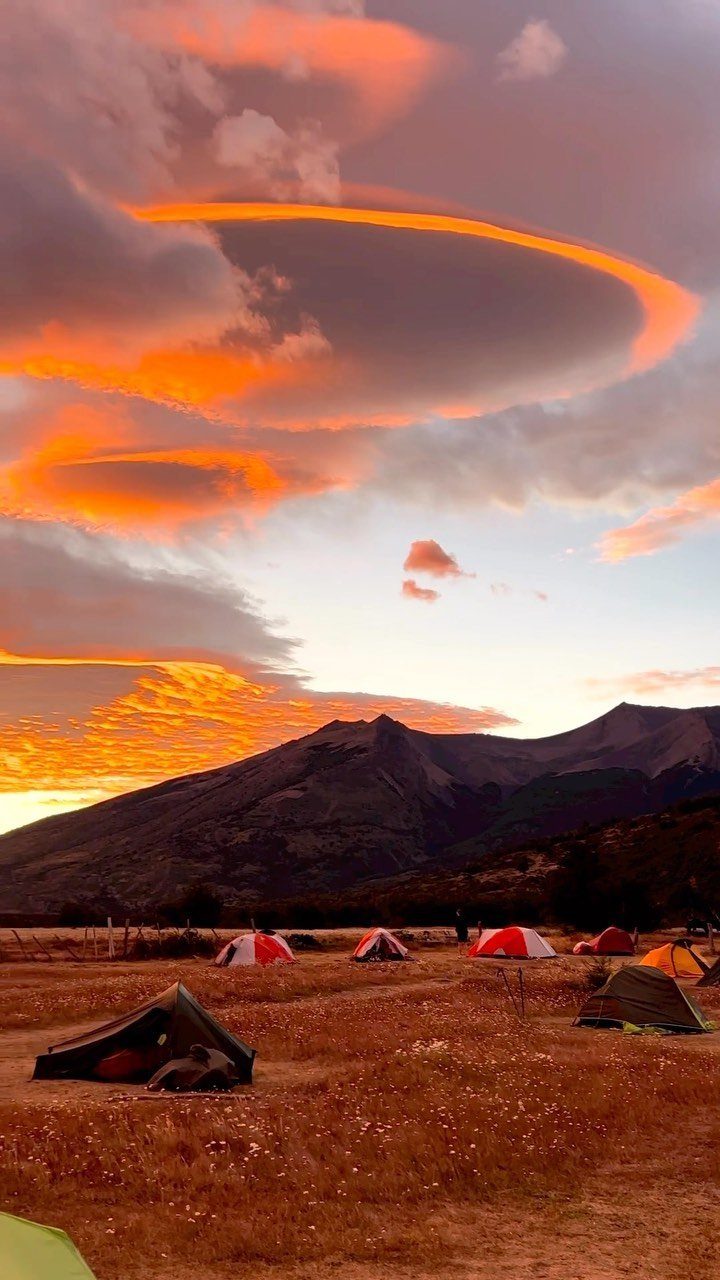 Camping tents in a grassy field before a mountain range under sunset clouds.