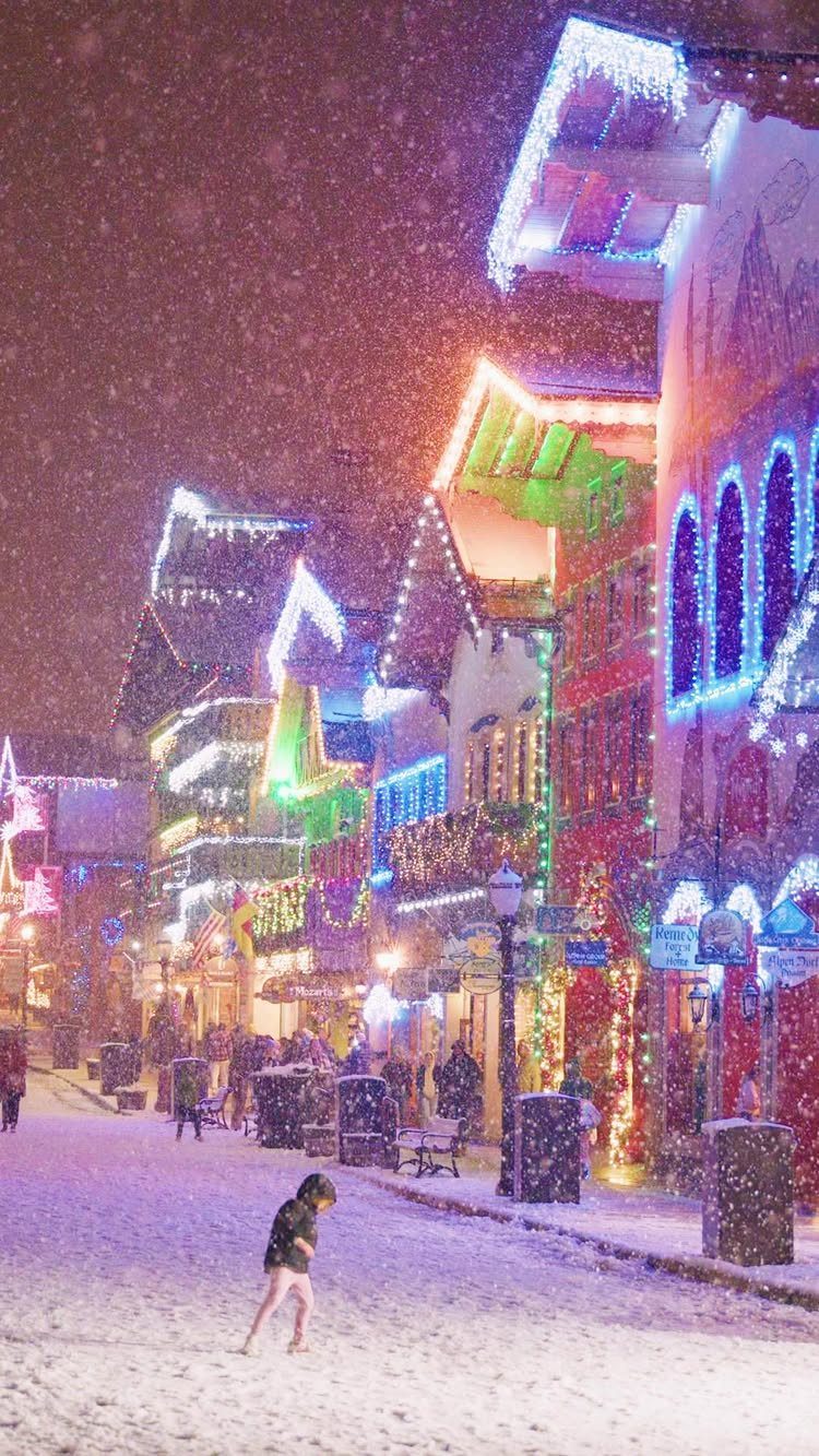 14 Christmas Places to Visit for Holiday Lights and Markets 2 Snowy village street with traditional buildings decorated in colorful festive holiday lights.