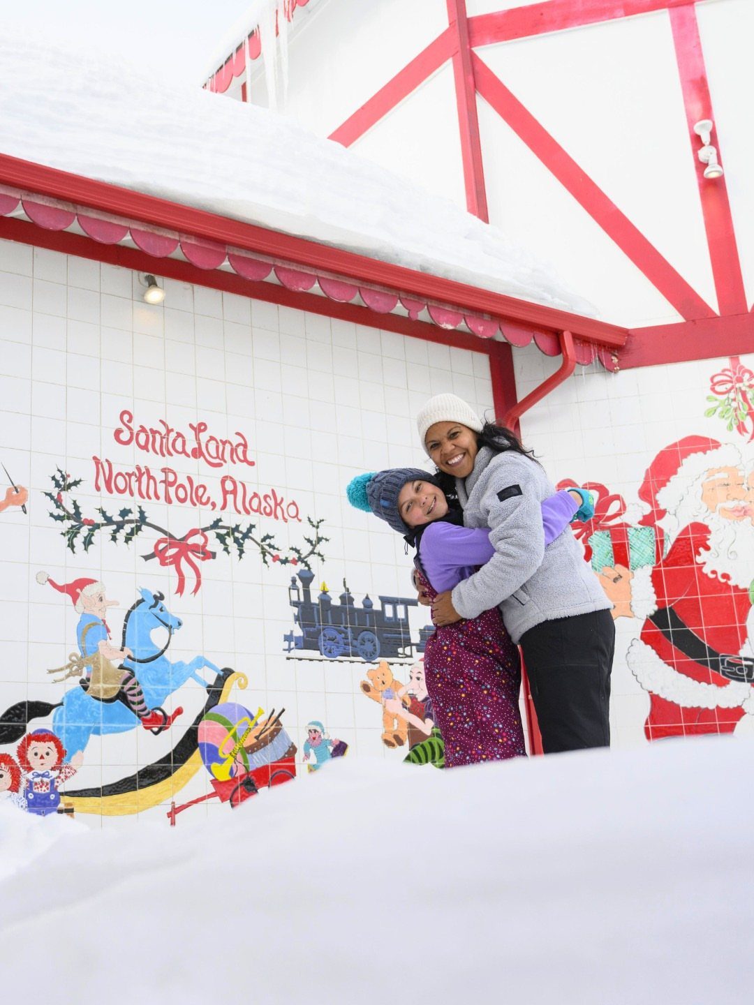 14 Christmas Places to Visit for Holiday Lights and Markets 4 Two women hugging in front of a Santa Land North Pole Alaska mural.