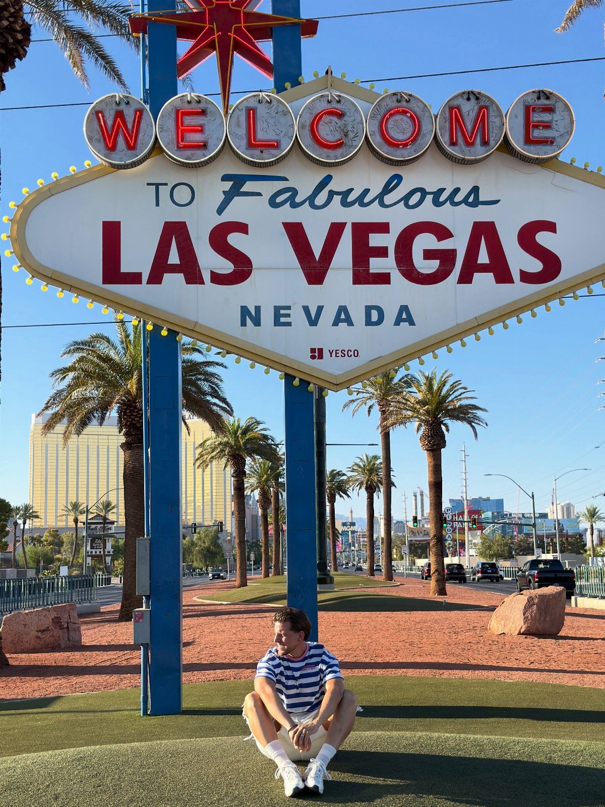 12 Cities to Visit in the U.S. and Abroad for 2026 7 Man sitting under the Welcome to Fabulous Las Vegas sign with palm trees behind.