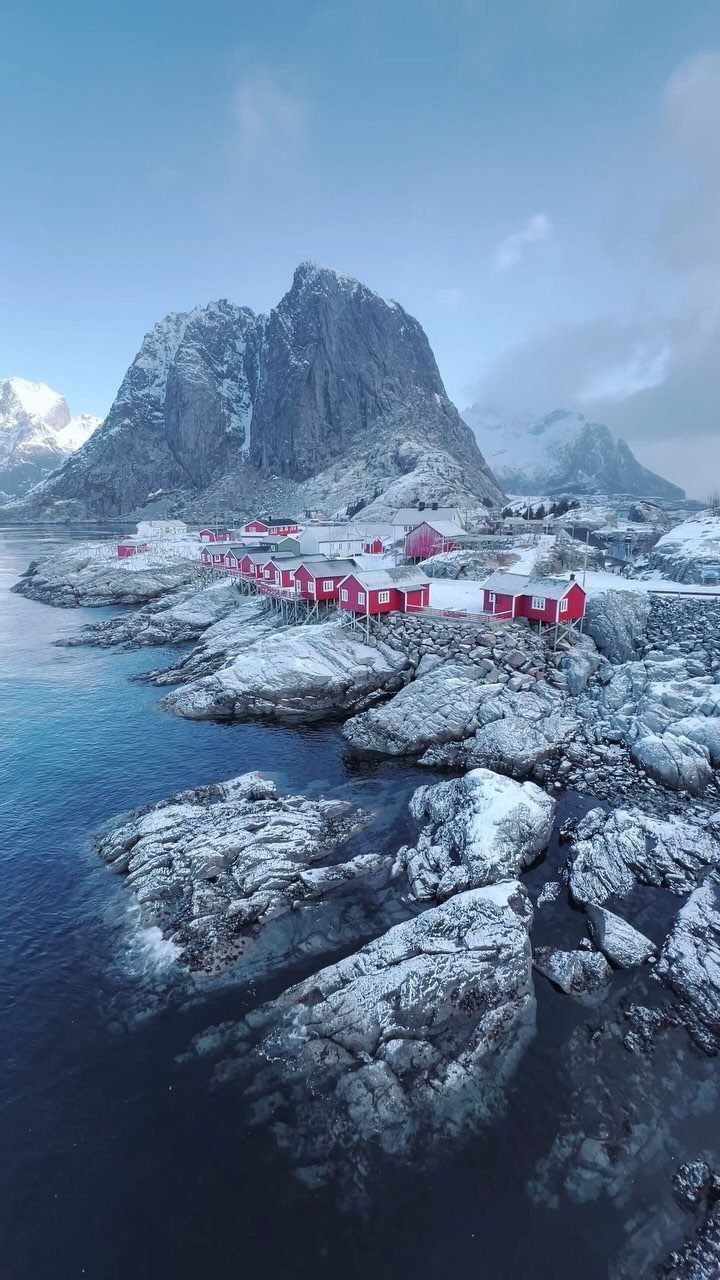 Red wooden cabins on a snowy rocky coast with a towering mountain peak.