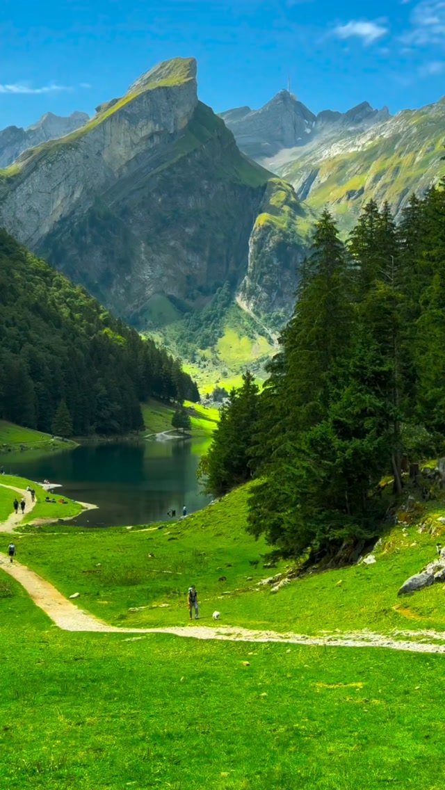Green valley with a dirt path and lake beneath towering rocky mountains.