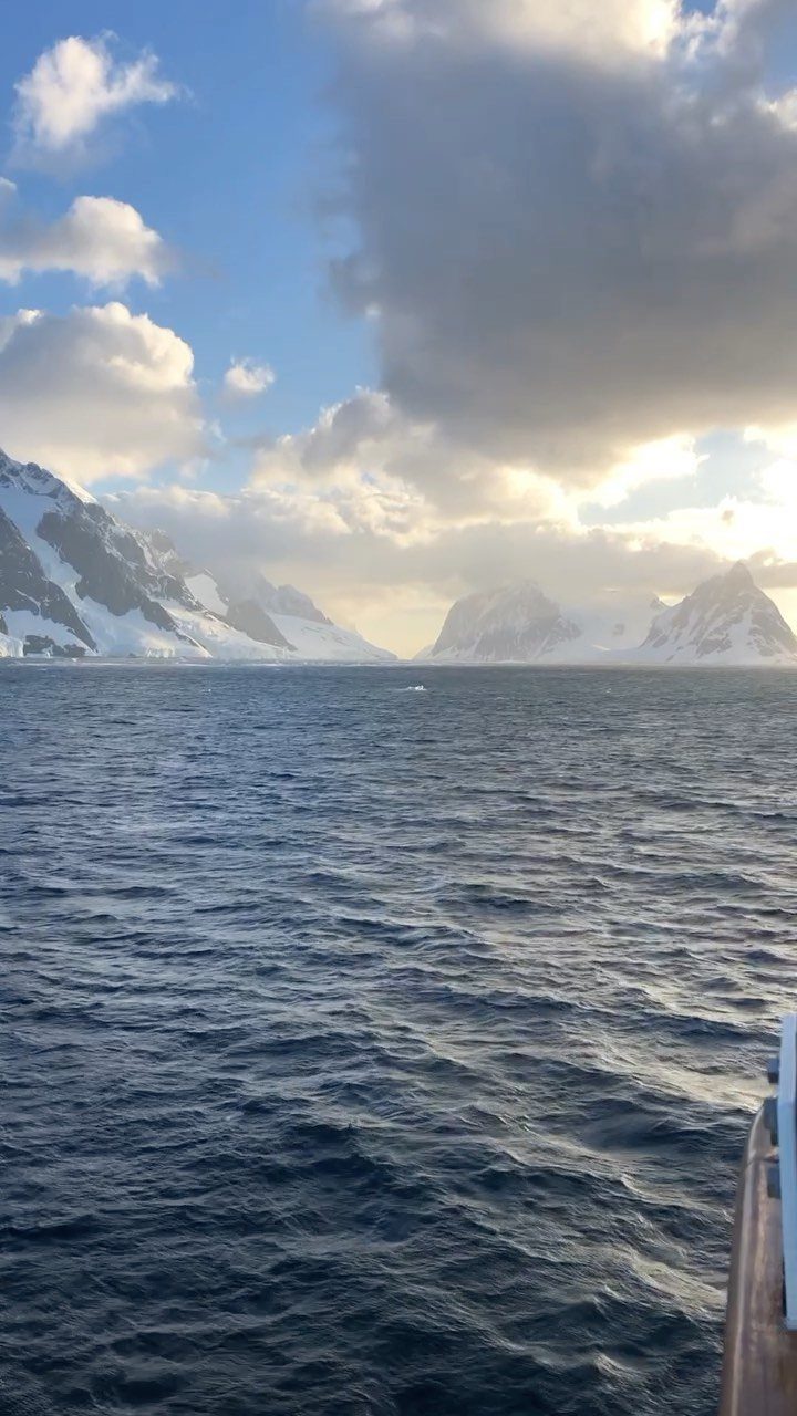 Snow-covered mountains and glaciers bordering a dark blue ocean under a cloudy sky.