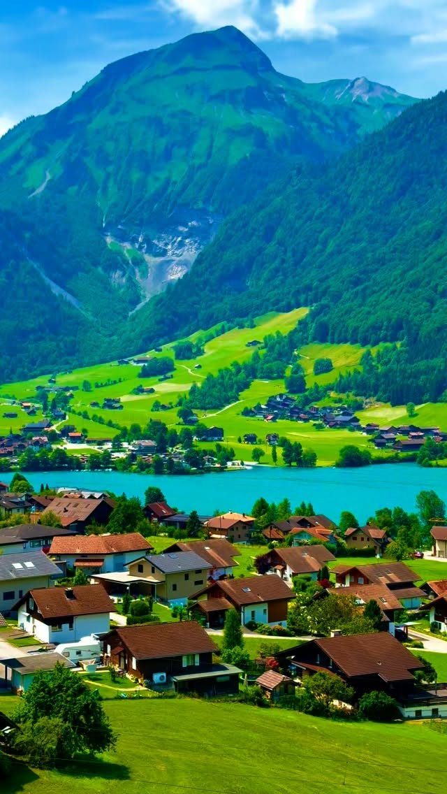 Green alpine village with brown houses beside a turquoise lake and steep mountains.