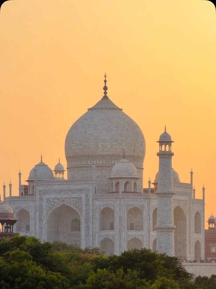 Taj Mahal white marble mausoleum under a warm orange sunset sky with greenery.