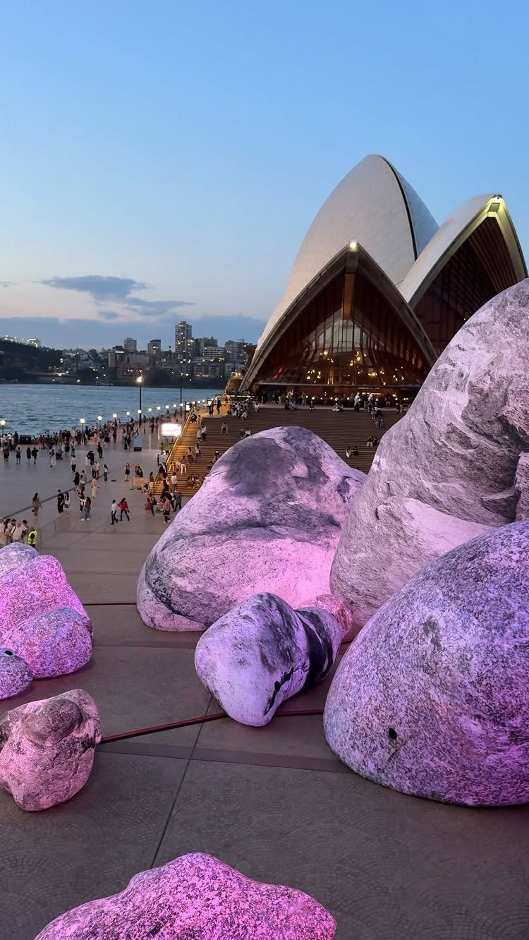 Sydney Opera House sails behind large purple-lit rocks on a harbor walkway.
