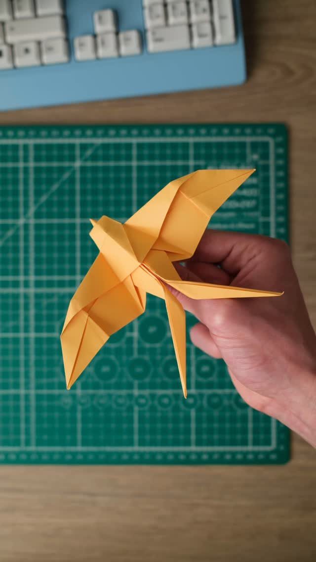 Yellow origami bird held by a hand over a green grid cutting mat.