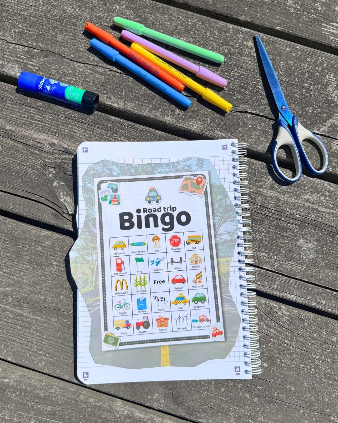 Road Trip Bingo card on a notebook with colorful markers and scissors on wood.
