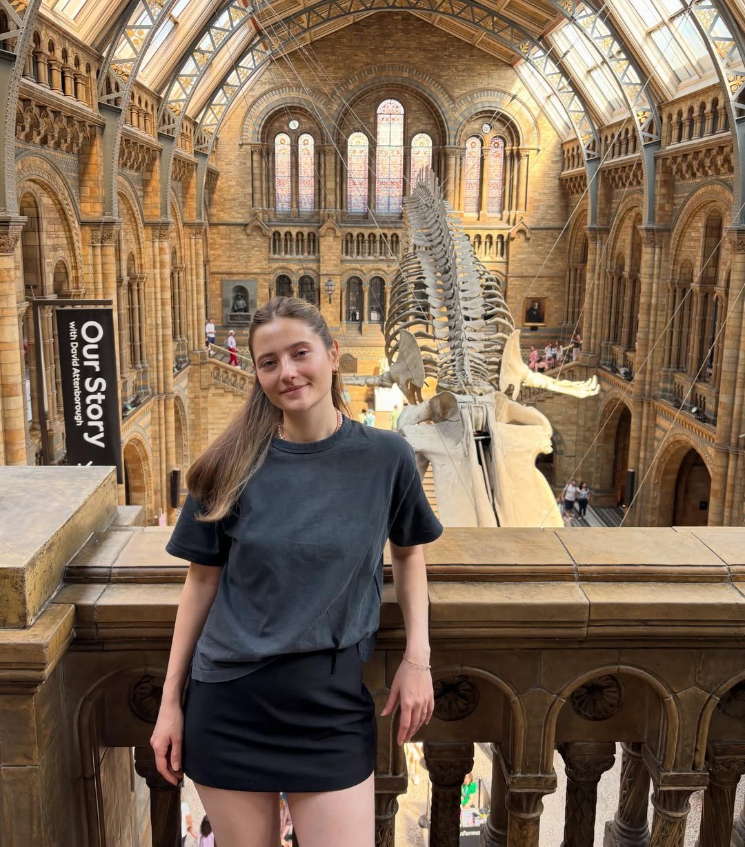 Young woman posing in a grand hall with a dinosaur skeleton behind her.