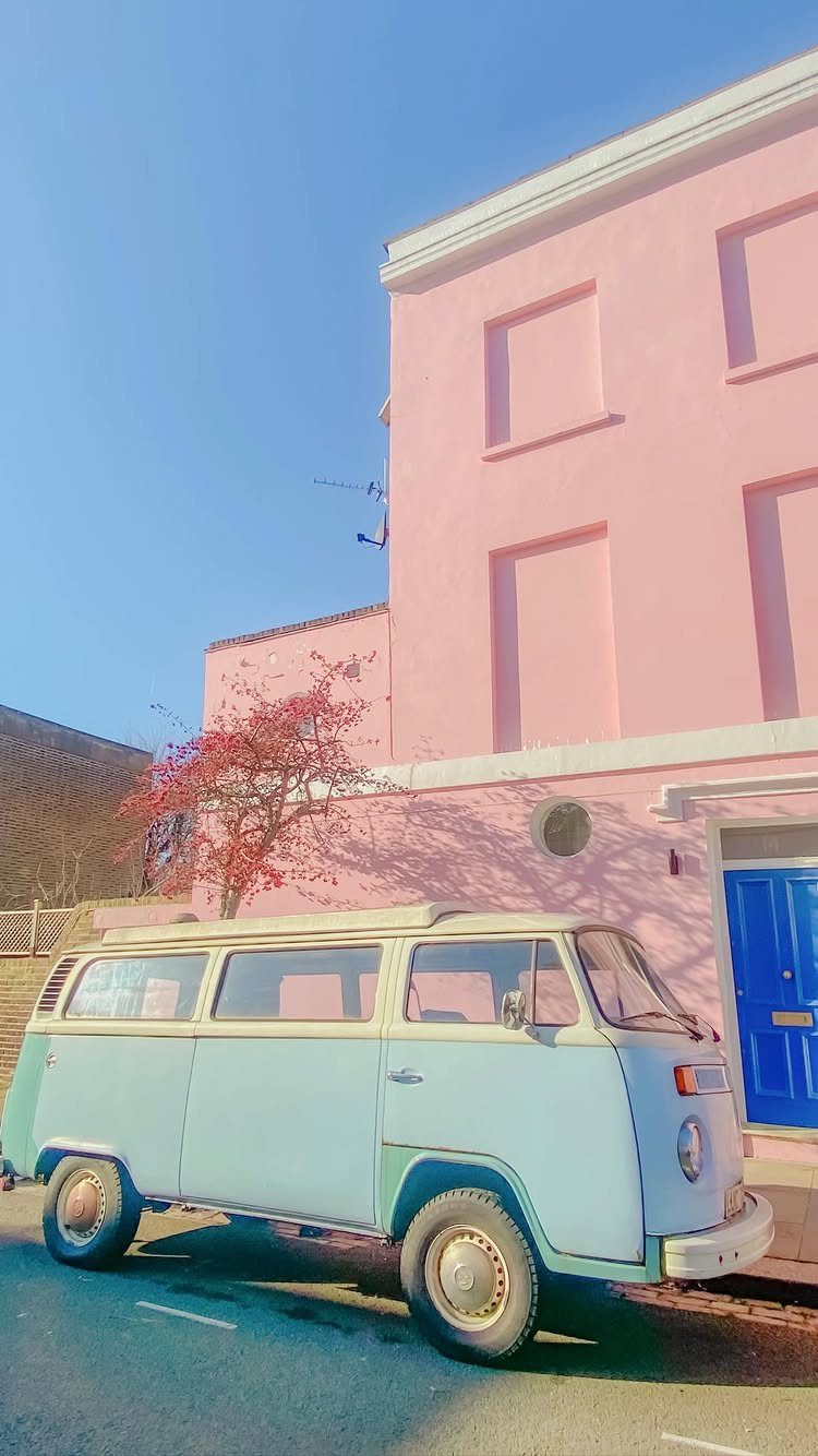 Light blue vintage camper van parked before a bright pink house with blue door.