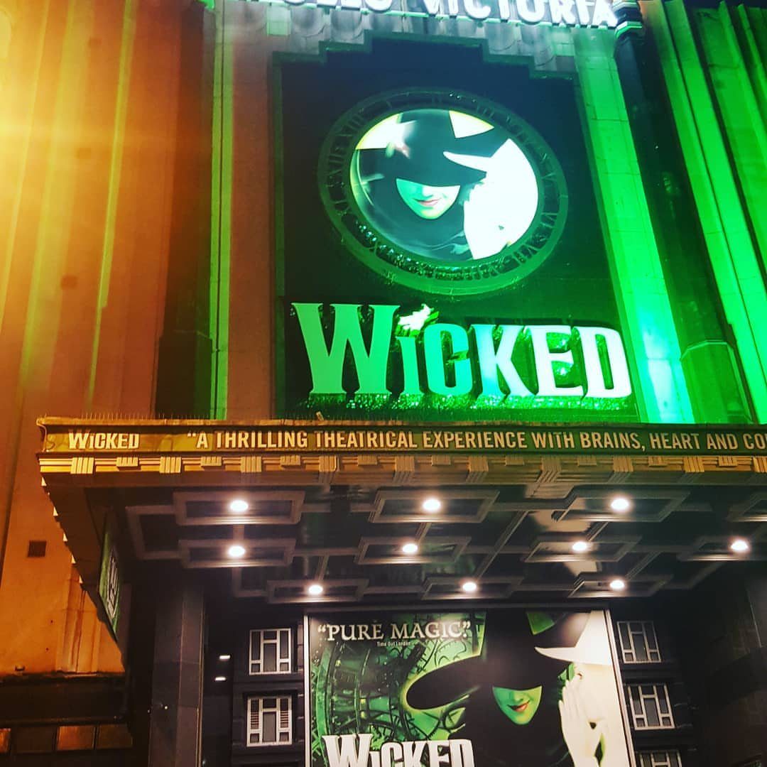 Wicked theater exterior with glowing green signage and backlit character portraits at night