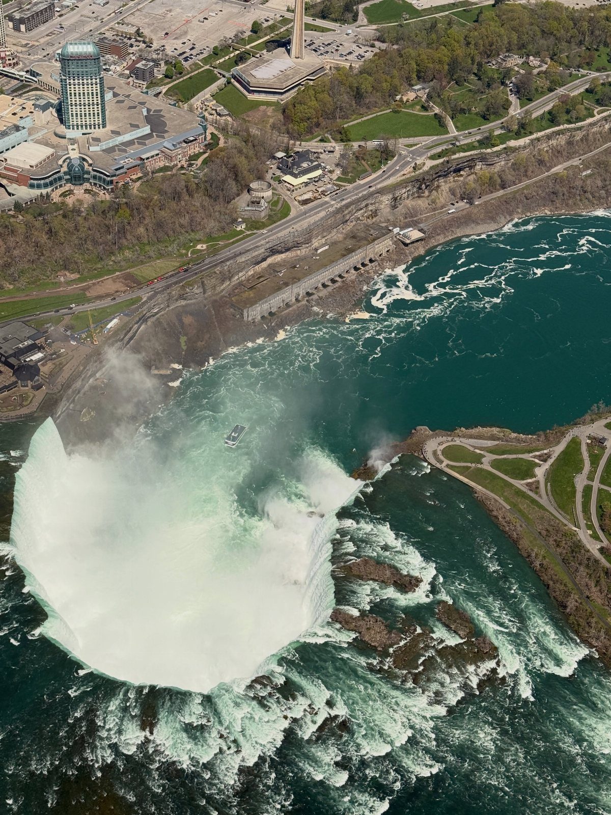 Horseshoe-shaped waterfall with white rushing water flowing into a turquoise river near city buildings.