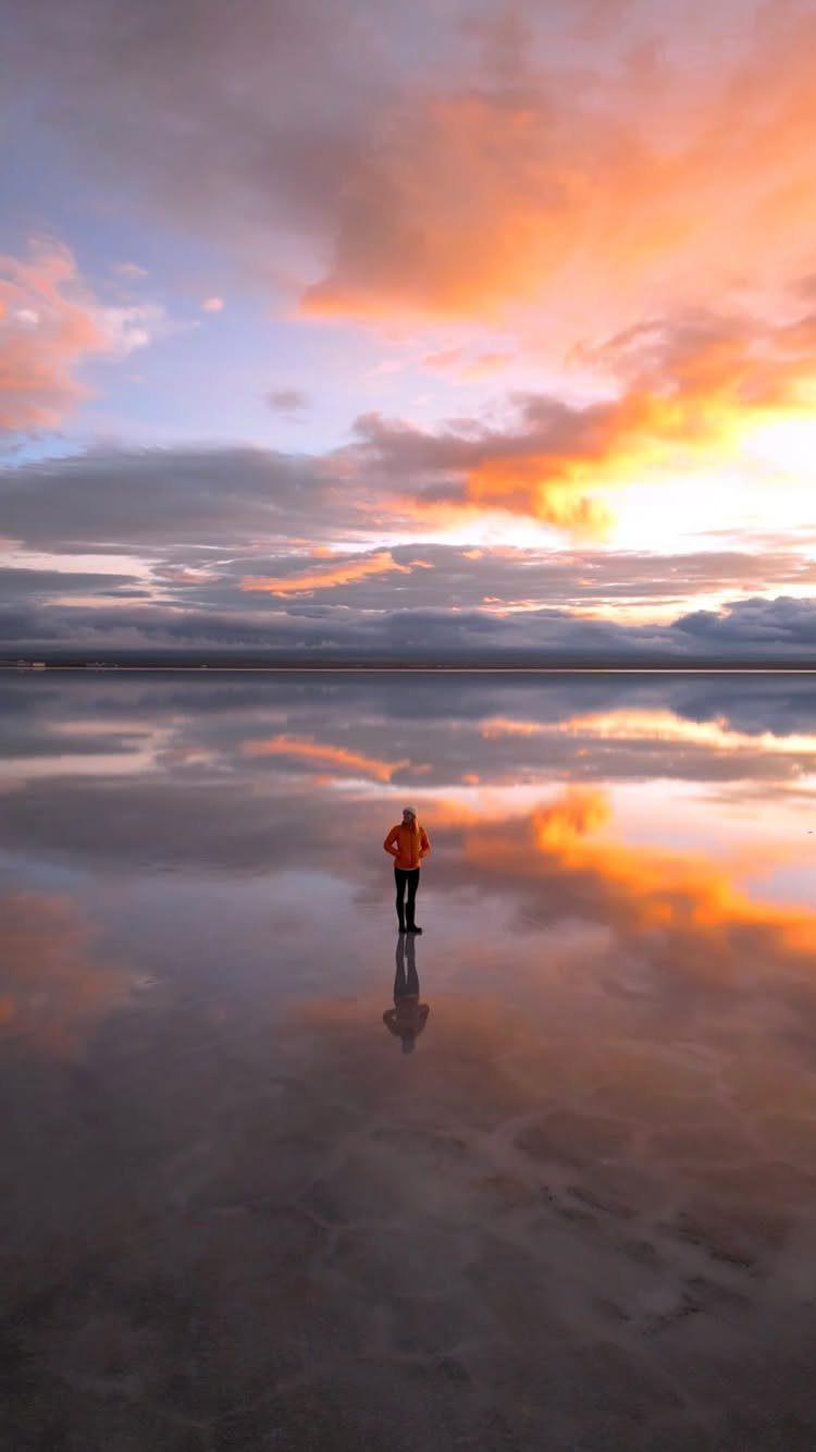 Person in orange jacket standing on reflective salt flats under a sunset sky