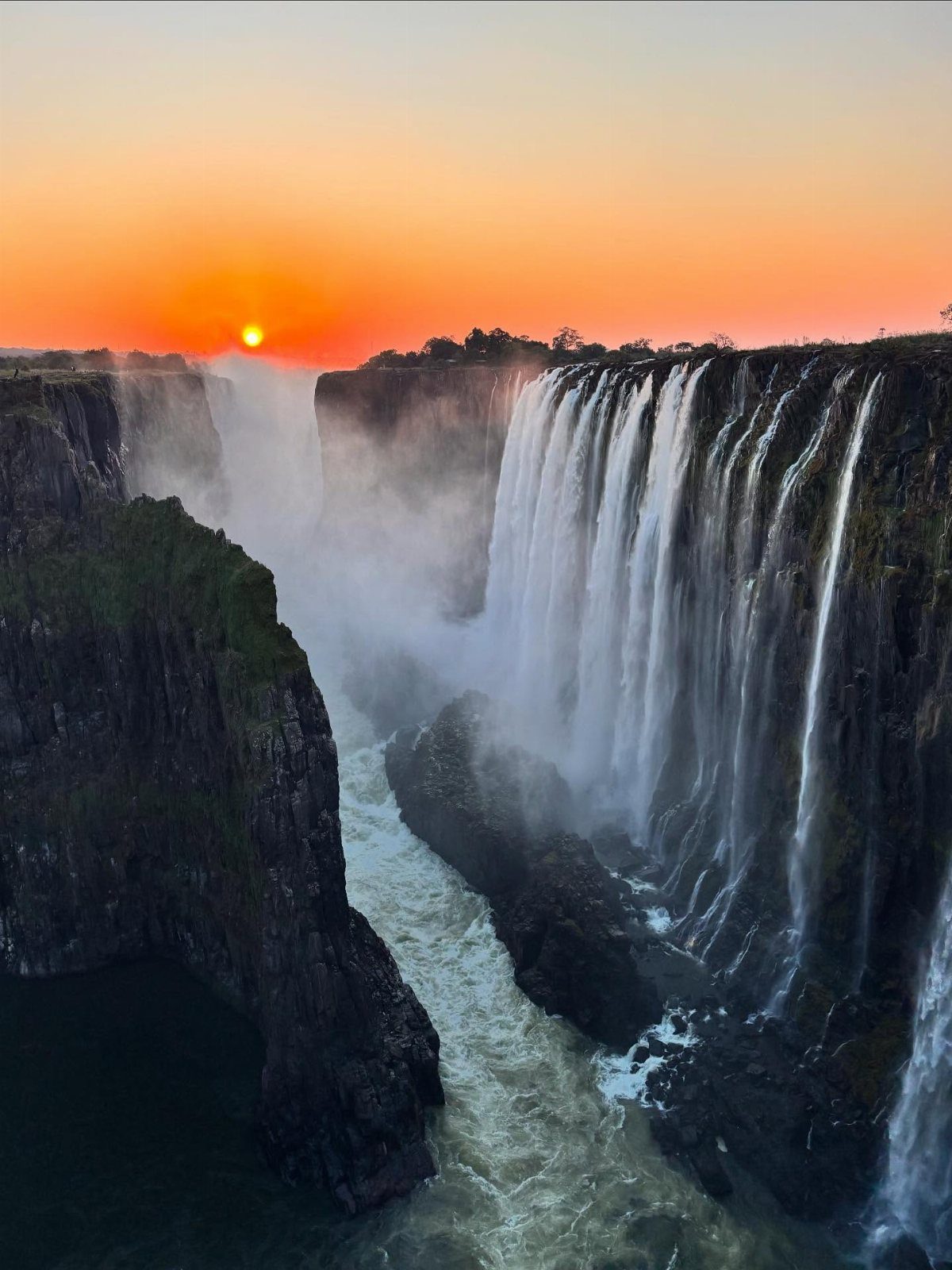 Massive waterfall cascading over dark cliffs under a vibrant orange sunset sky.