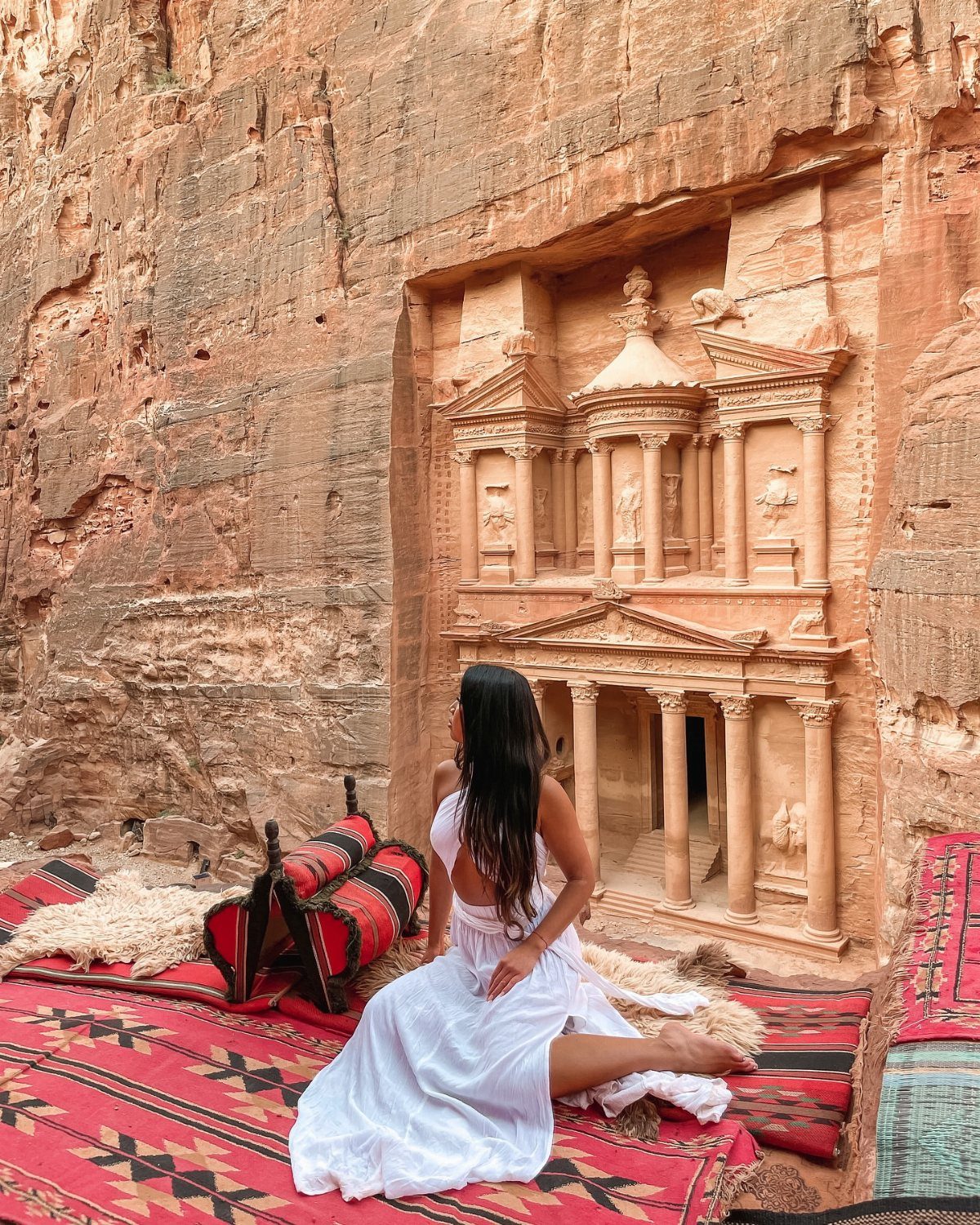 Woman in white dress sitting before the Treasury carved into red rock walls.