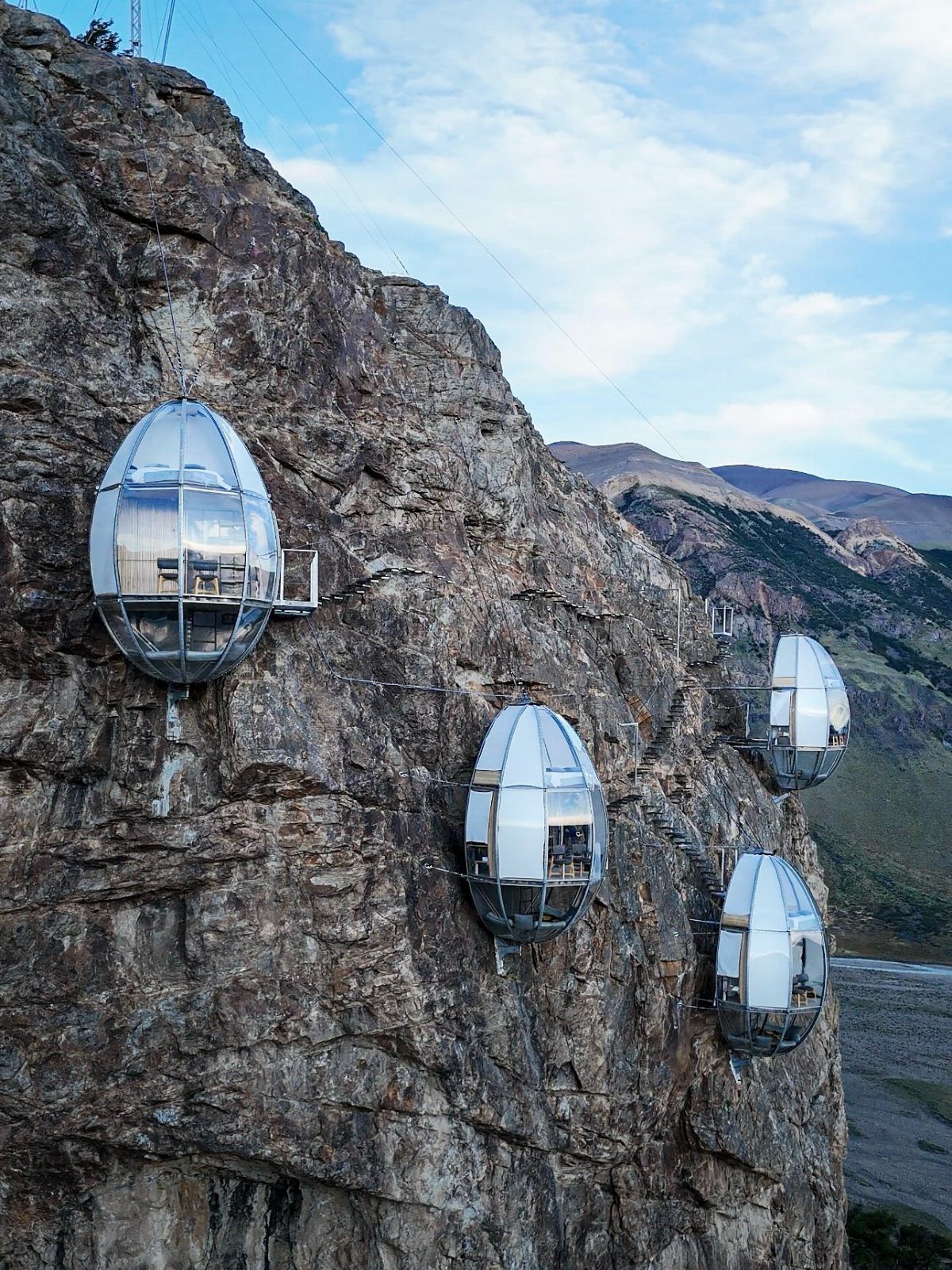 Futuristic glass pods attached to a rocky cliff with mountains in the background
