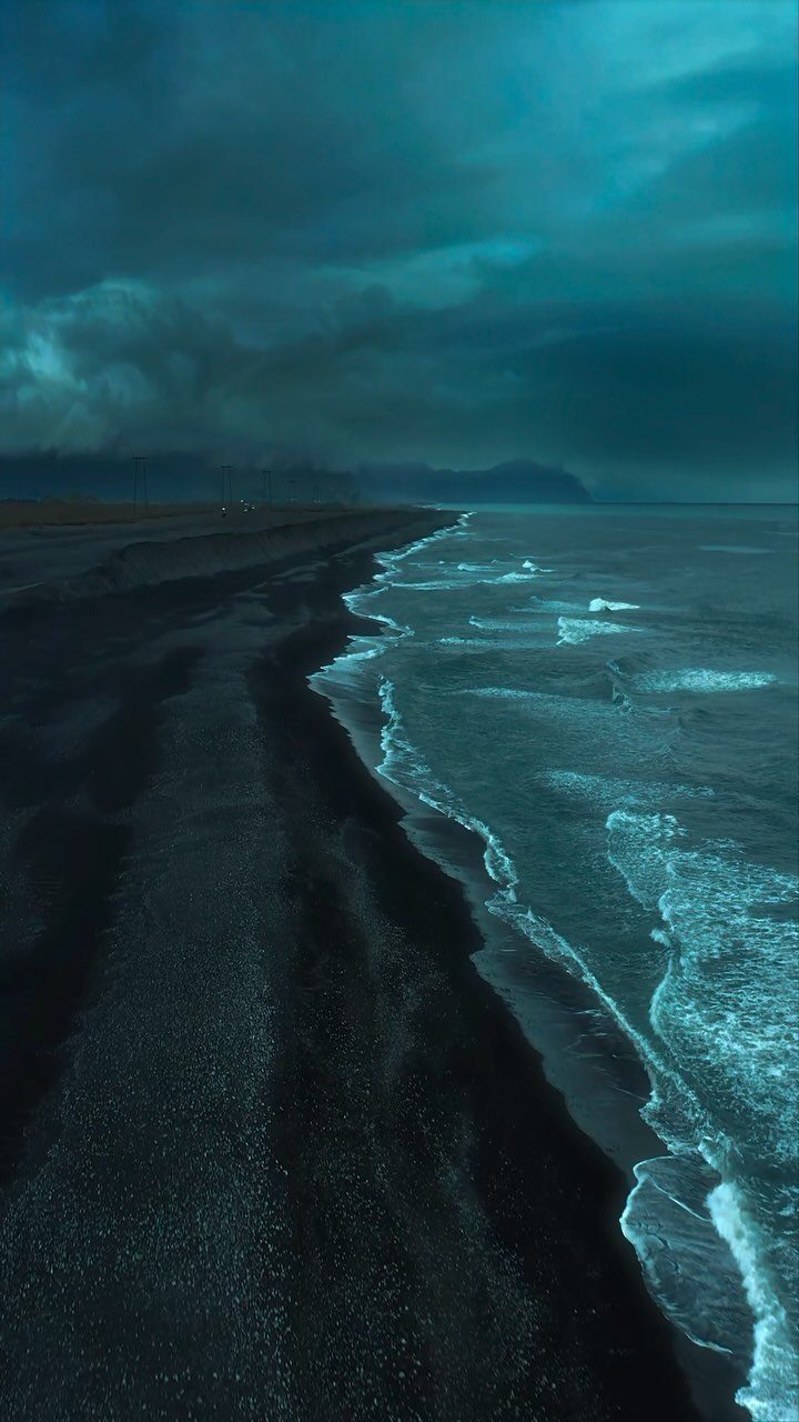 Black sand beach with turquoise waves under a dark, moody teal sky.