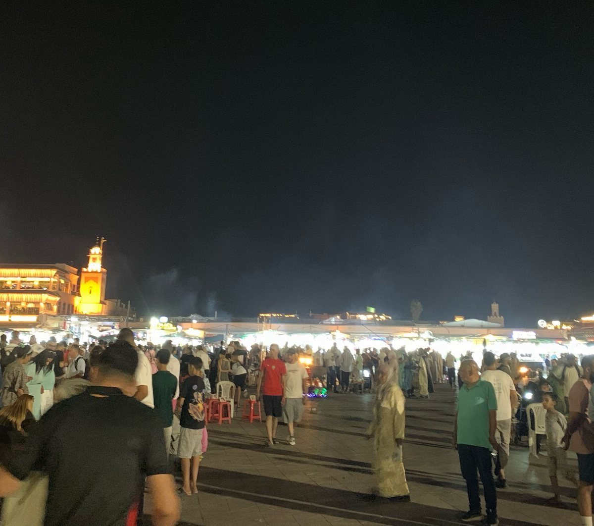 Crowded night market in a city square with lit stalls and distant tower.
