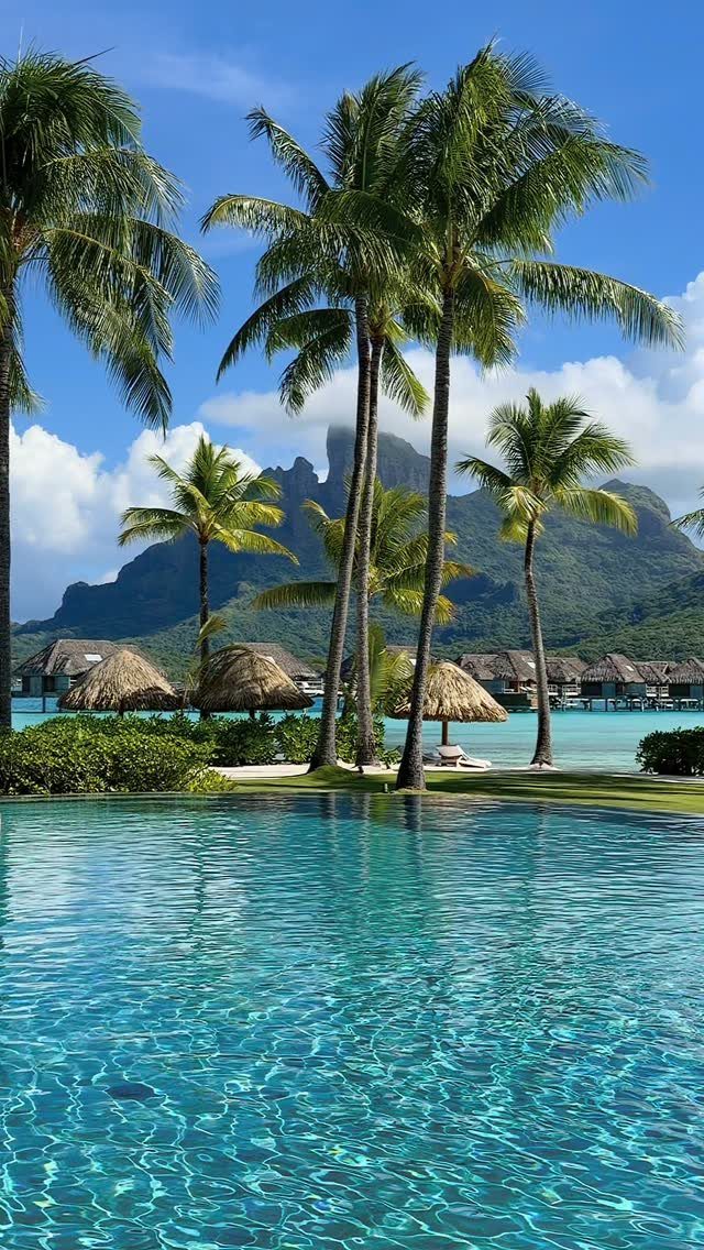 Tropical resort pool with palm trees and overwater bungalows before a mountain peak.