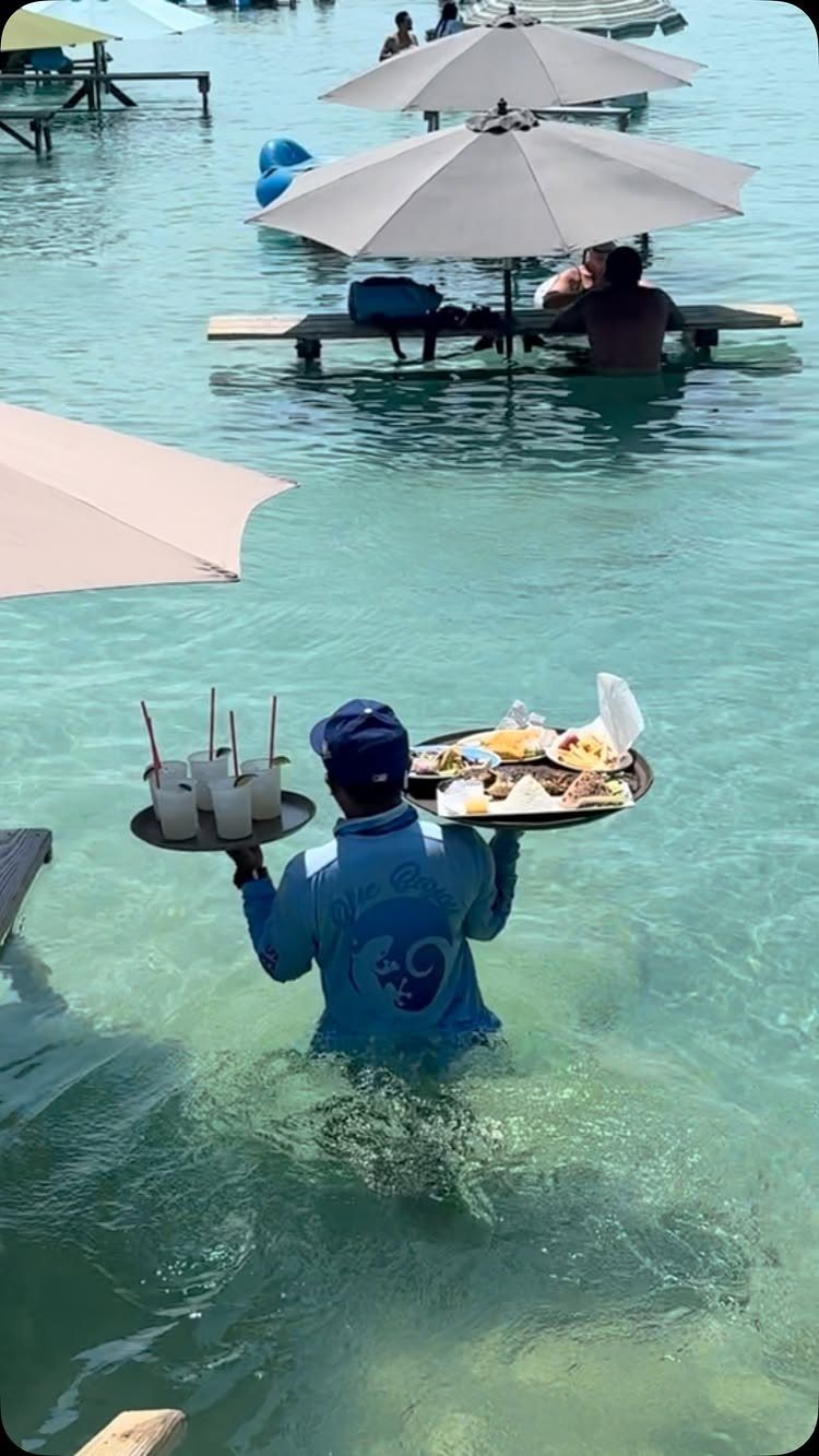 12 Places Like Bora Bora for Island Views and Lagoons 10 Server wading through turquoise lagoon water carrying food trays toward beach umbrellas.