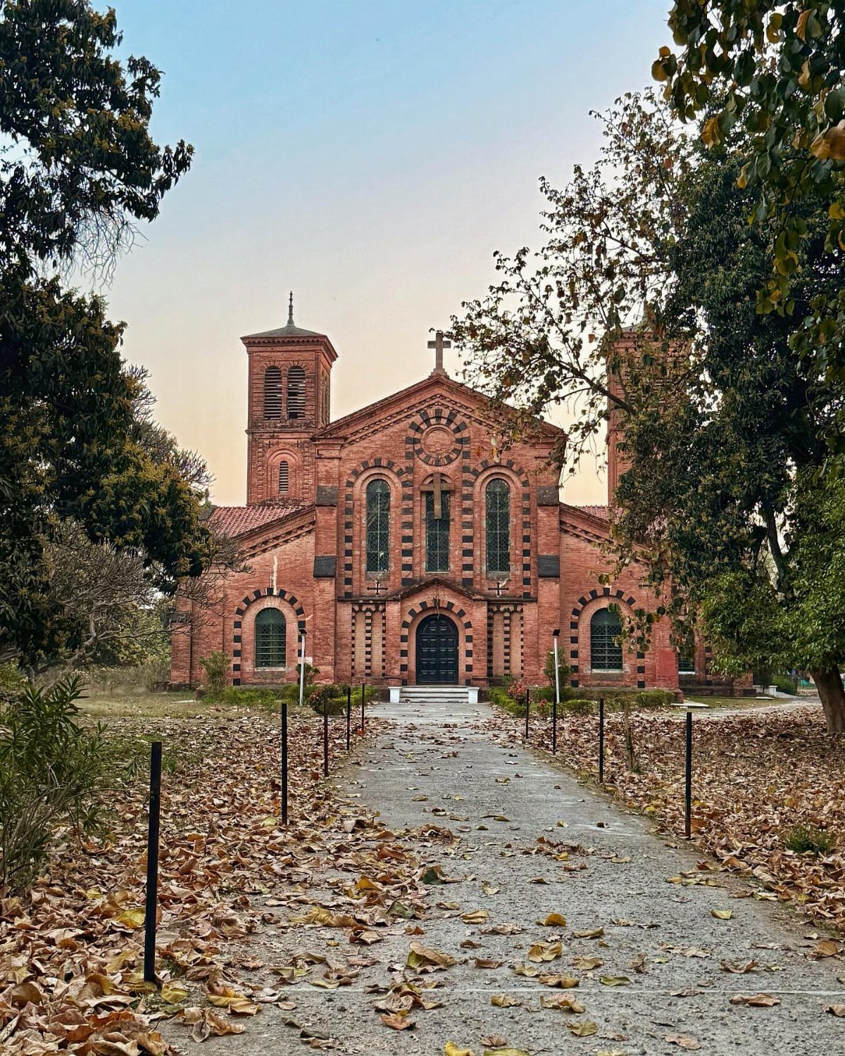 Red brick church with towers and a cross viewed from a leaf-covered path.