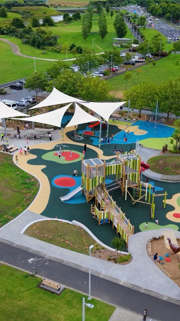 Modern outdoor playground with colorful flooring, wooden climbing structures, and white shade sails.
