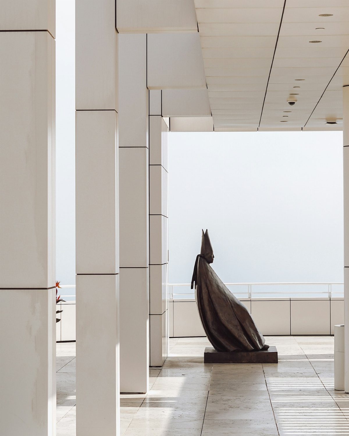 Bronze cloaked figure sculpture standing among white modern columns in an outdoor gallery.